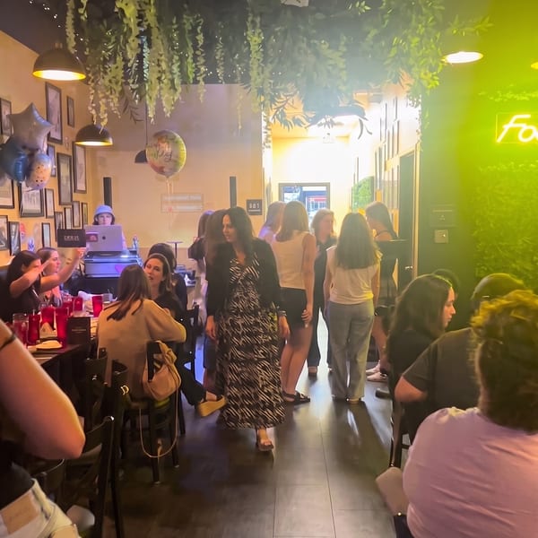 A lively restaurant interior with patrons dining, a DJ, and hanging greenery under warm lighting.