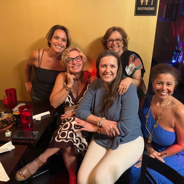 Five smiling women pose together at a restaurant table with drinks and food, under disco lights.