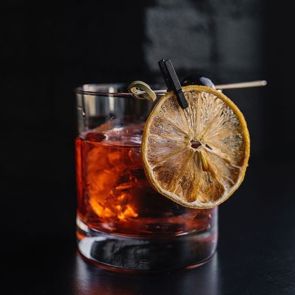 A cocktail in a rocks glass with ice, garnished with a dried orange slice and olive.