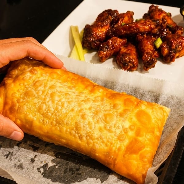Hand holding a large fried pastry next to a plate of glazed chicken wings with dipping sauce.