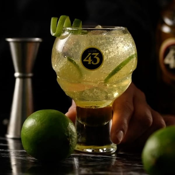 A hand holds a Licor 43 cocktail with lime slices and a lime peel garnish, surrounded by whole limes.