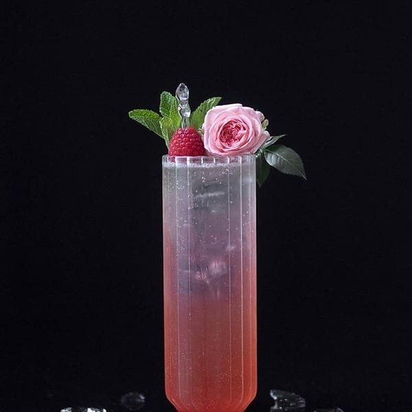 Pink cocktail in a tall ribbed glass, garnished with a raspberry, mint, and a pink rose.