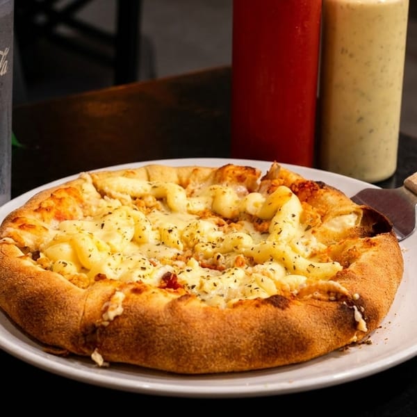 Personal mac and cheese pizza with a pizza server, Coca-Cola, and condiments on a dark table.